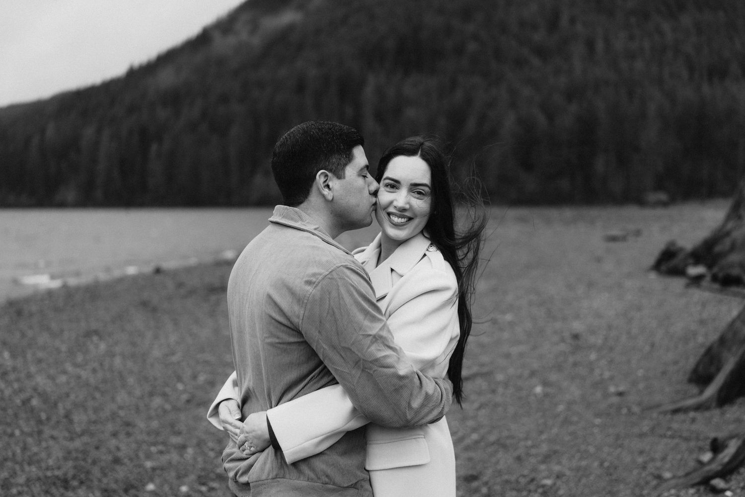 A couple embraces on a rocky beach with mountains and forest creating a dramatic backdrop.
