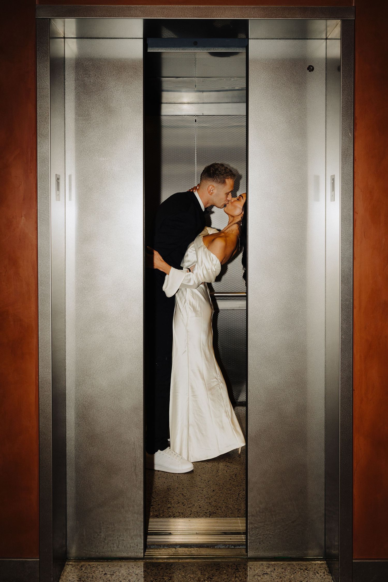 A couple shares an intimate kiss in an elevator wearing formal wedding attire.