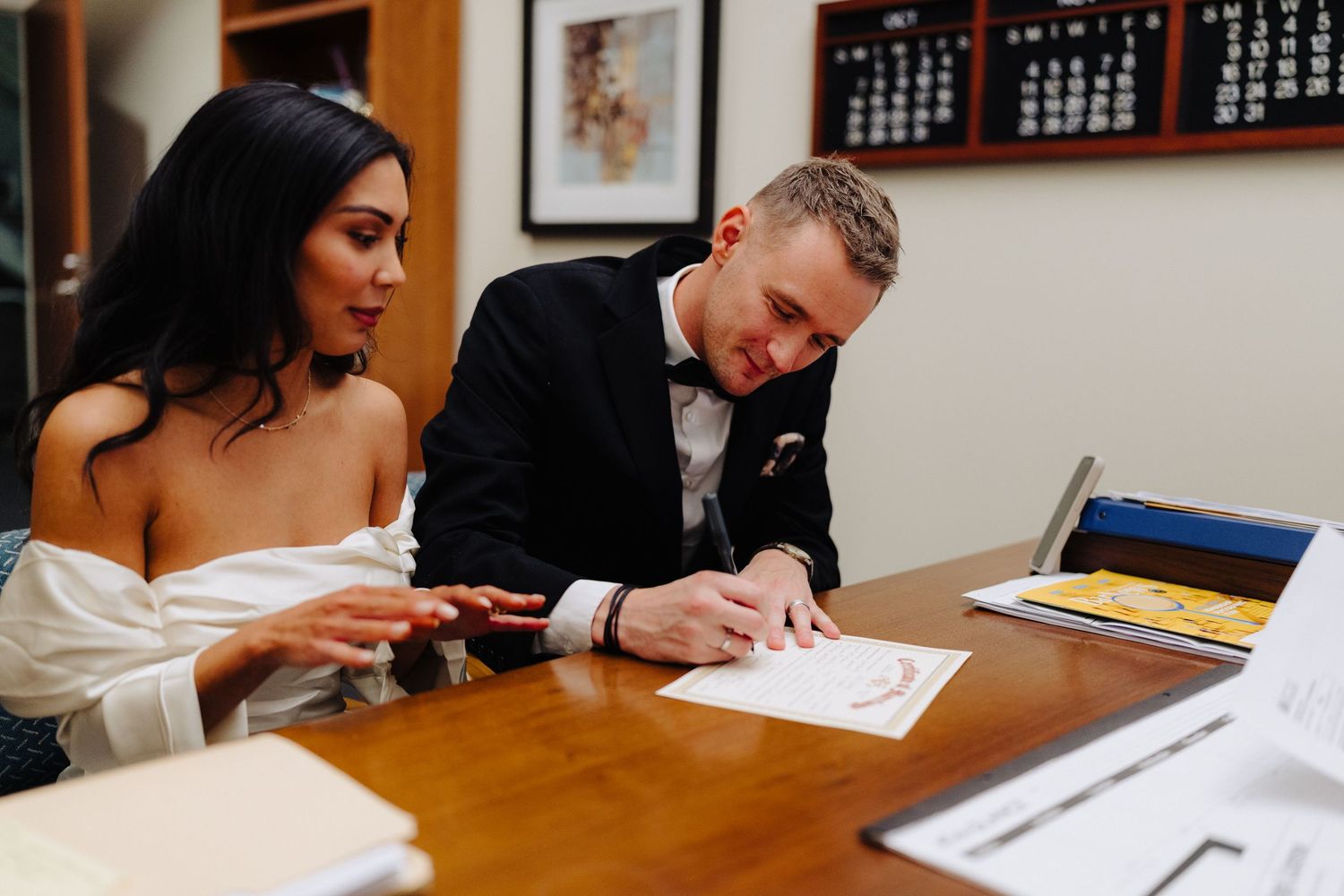 A couple signs documents at a wooden table during their wedding ceremony.