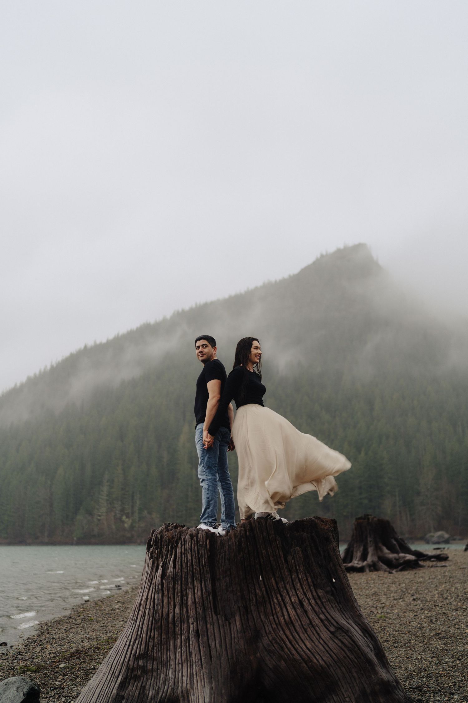 A couple stands atop a large tree stump by a misty mountain lake on a moody overcast day.