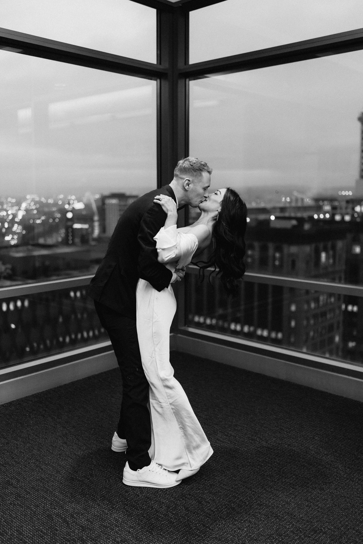 A romantic couple in black and white embrace and share a tender moment in a corner window overlooking a city skyline at night.