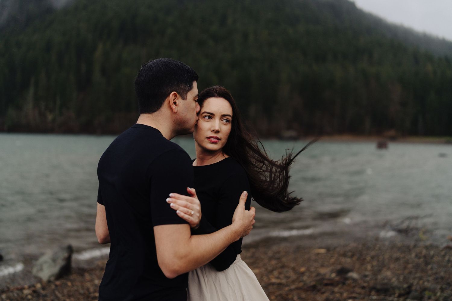 A romantic couple embraces on a moody shoreline with mountains and forest in the background.