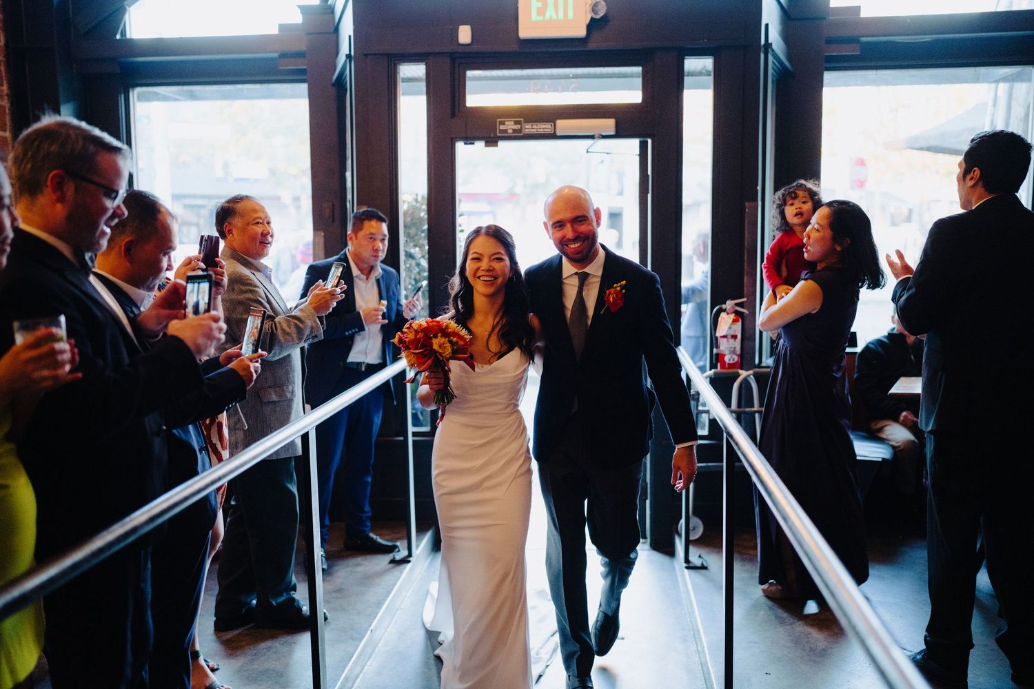 Bride and groom exit through a corridor lined with guests holding champagne glasses at an indoor venue.