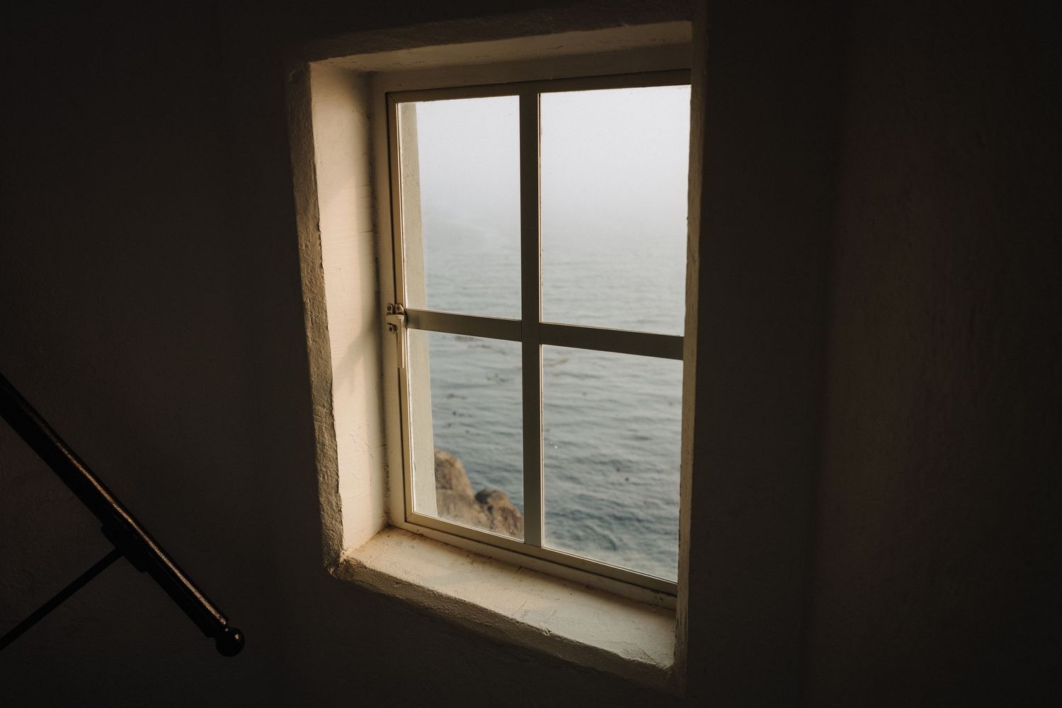 A window frame in a dark room overlooks the grey ocean waters on a moody day.