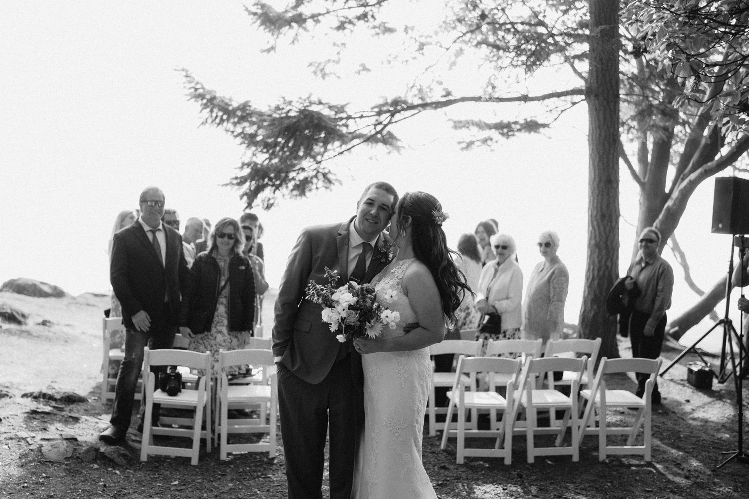 A romantic black and white wedding ceremony takes place outdoors under tall pine trees by the ocean.