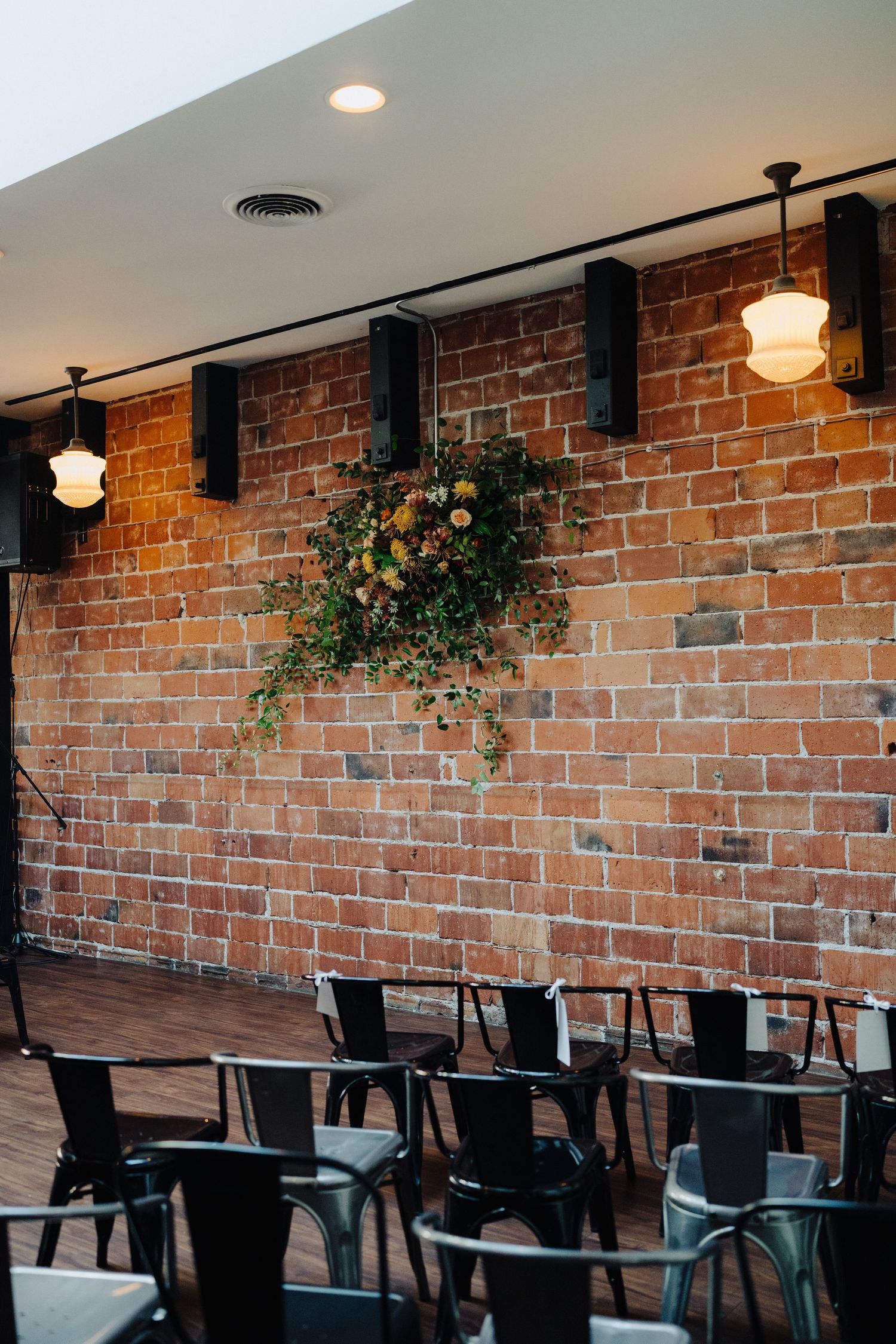 Industrial-chic restaurant interior with exposed brick walls, pendant lights, and black chairs arranged around tables.