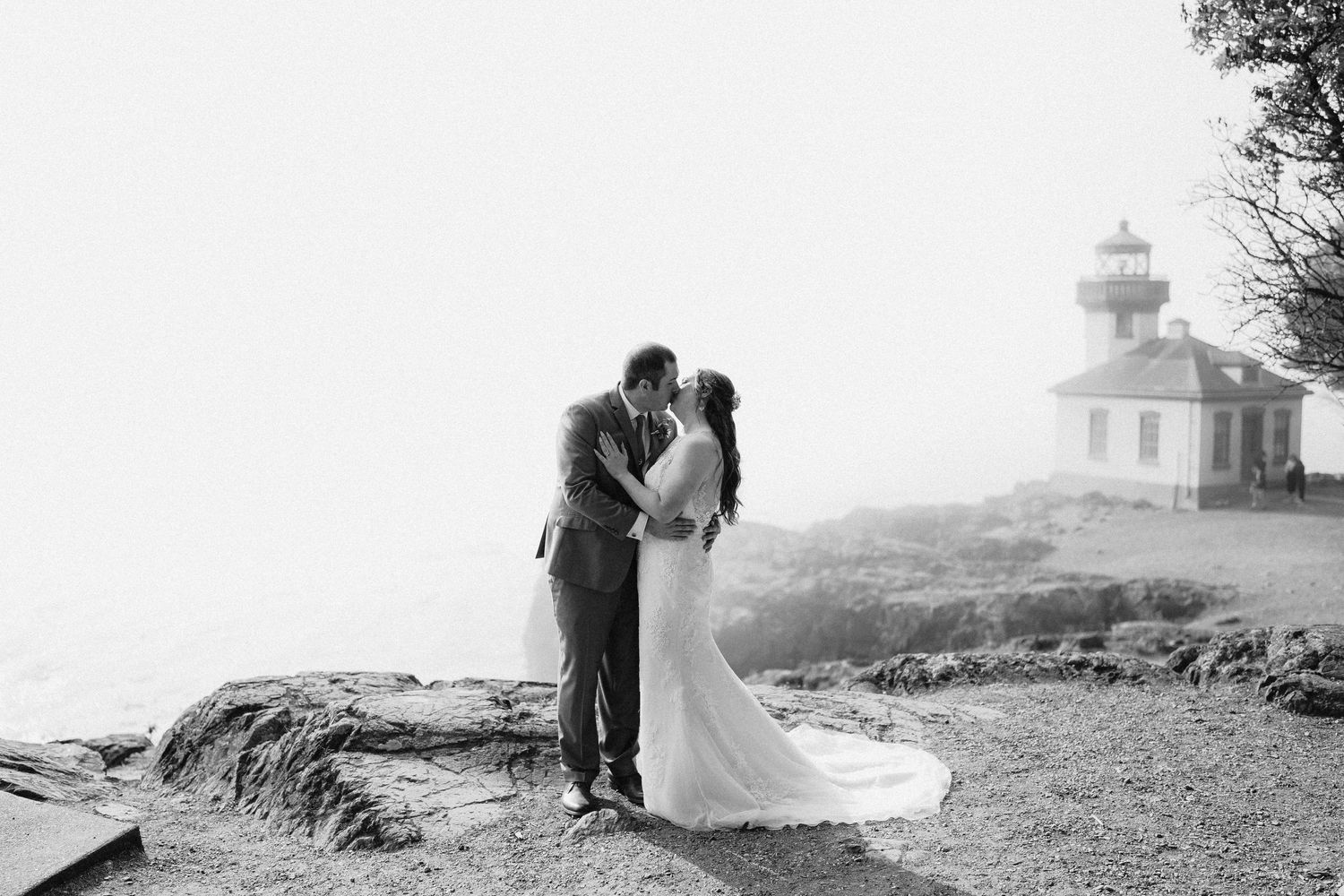 A romantic black and white wedding photo silhouette captured on a misty cliff overlooking a lighthouse in the distance.