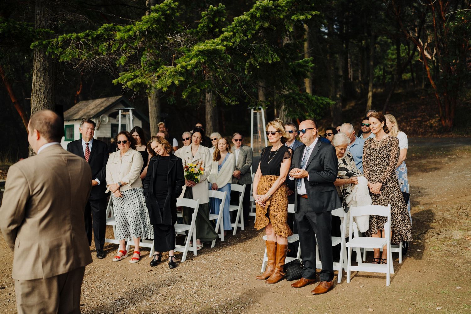 Wedding guests stand expectantly along a dirt path bordered by evergreen trees during an outdoor ceremony.