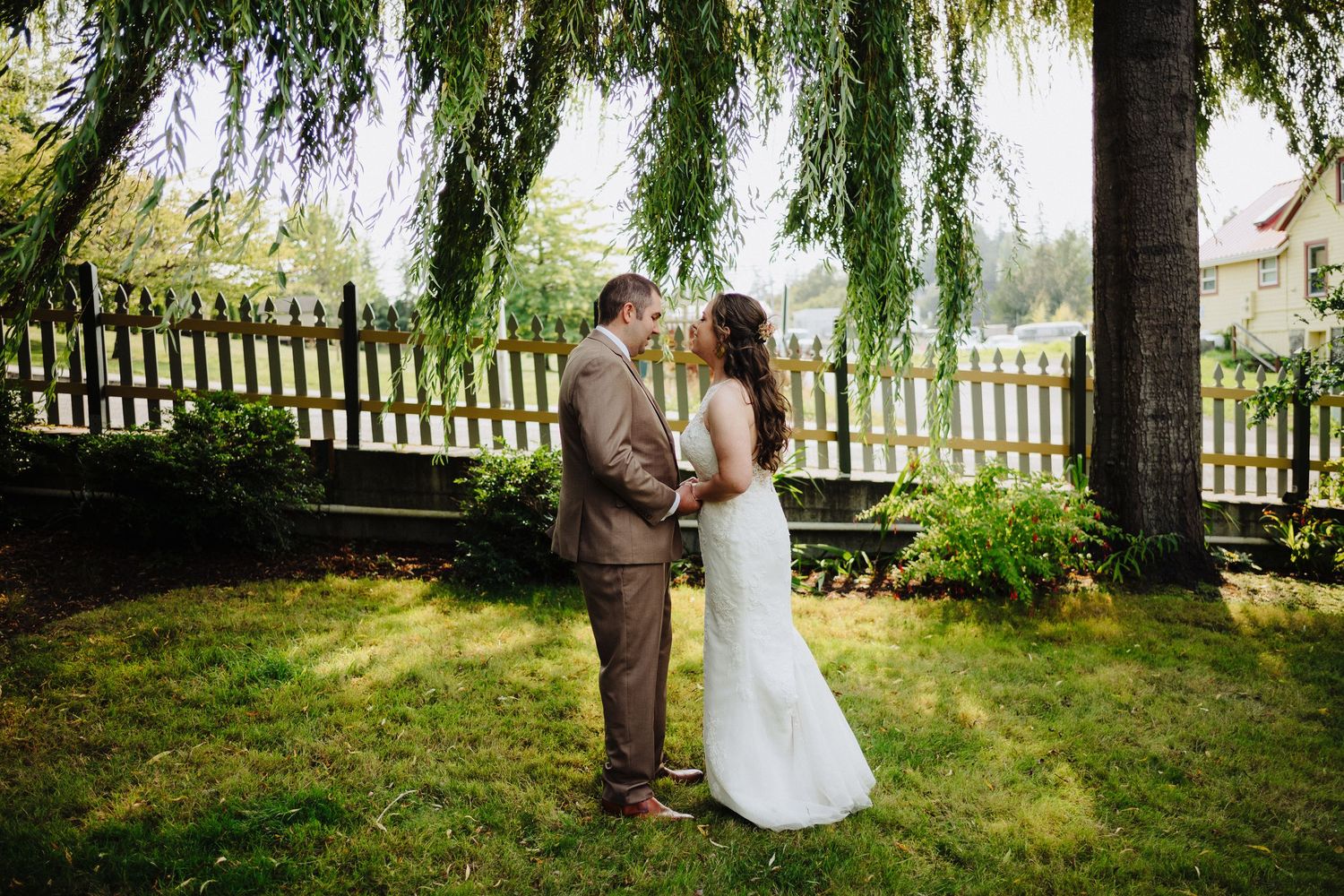 A couple embraces under a weeping willow tree during a garden wedding ceremony.