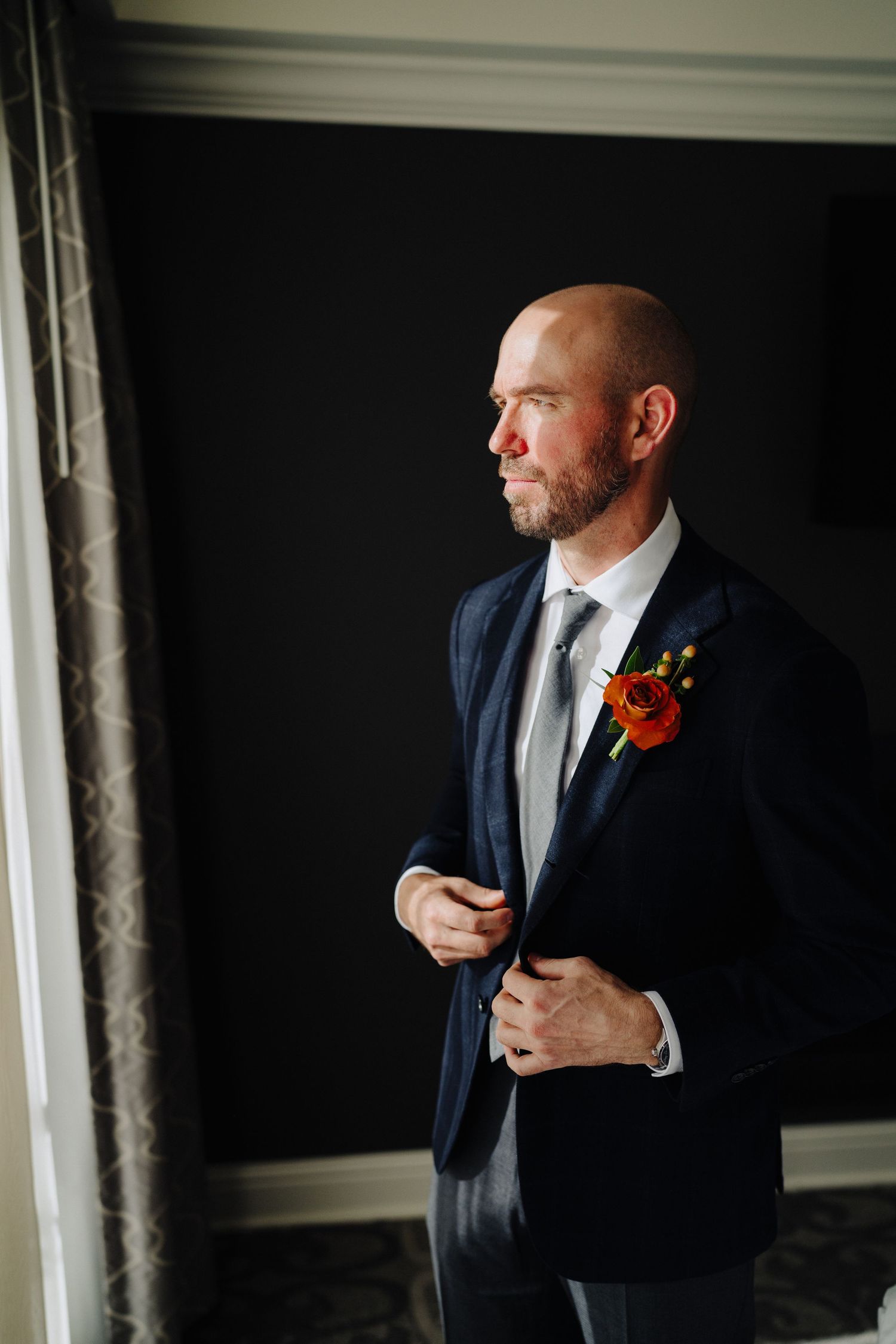 A groom in a navy suit with an orange boutonniere adjusts his jacket while standing near a window in dramatic lighting.