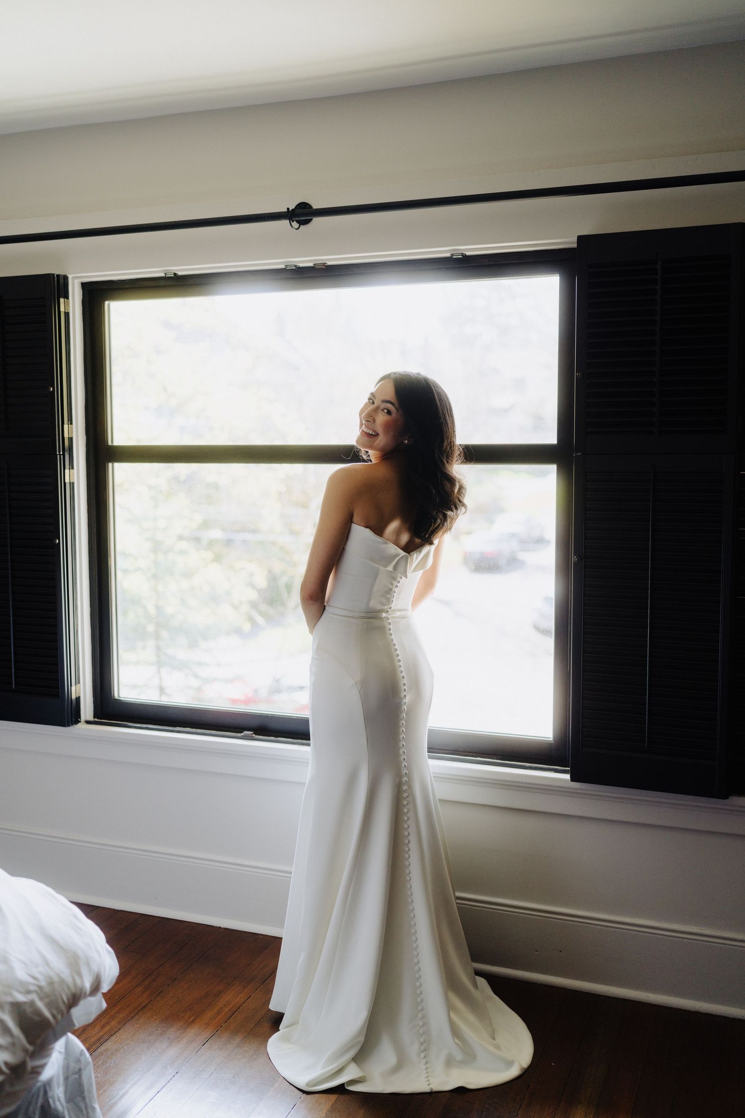 A person in a white wedding gown stands by a window in a sunlit room.