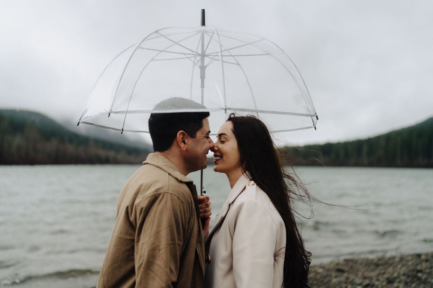 A couple shares a romantic moment under a clear umbrella by a misty lake with mountains in the distance.