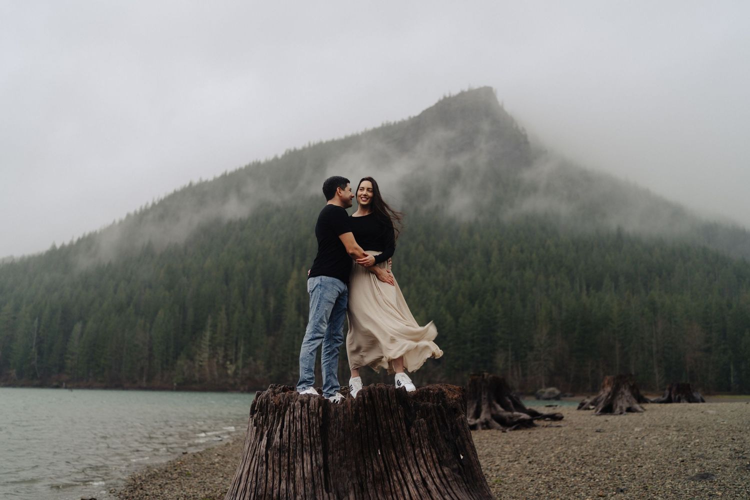 A couple stands embracing on a tree stump by a misty mountain lake surrounded by evergreen forest.