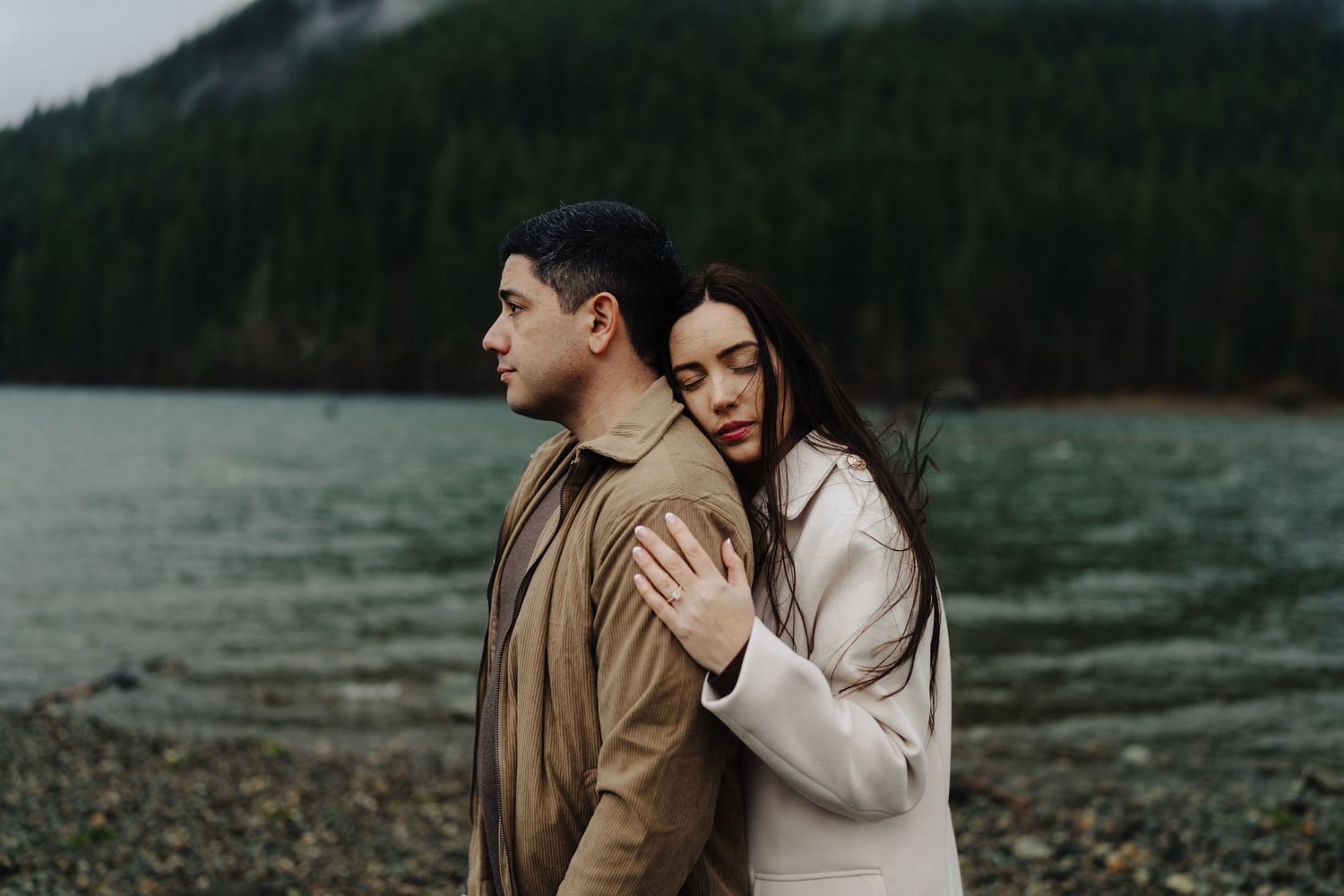 A couple embraces on a pebble beach shore with fog-covered mountains in the background.