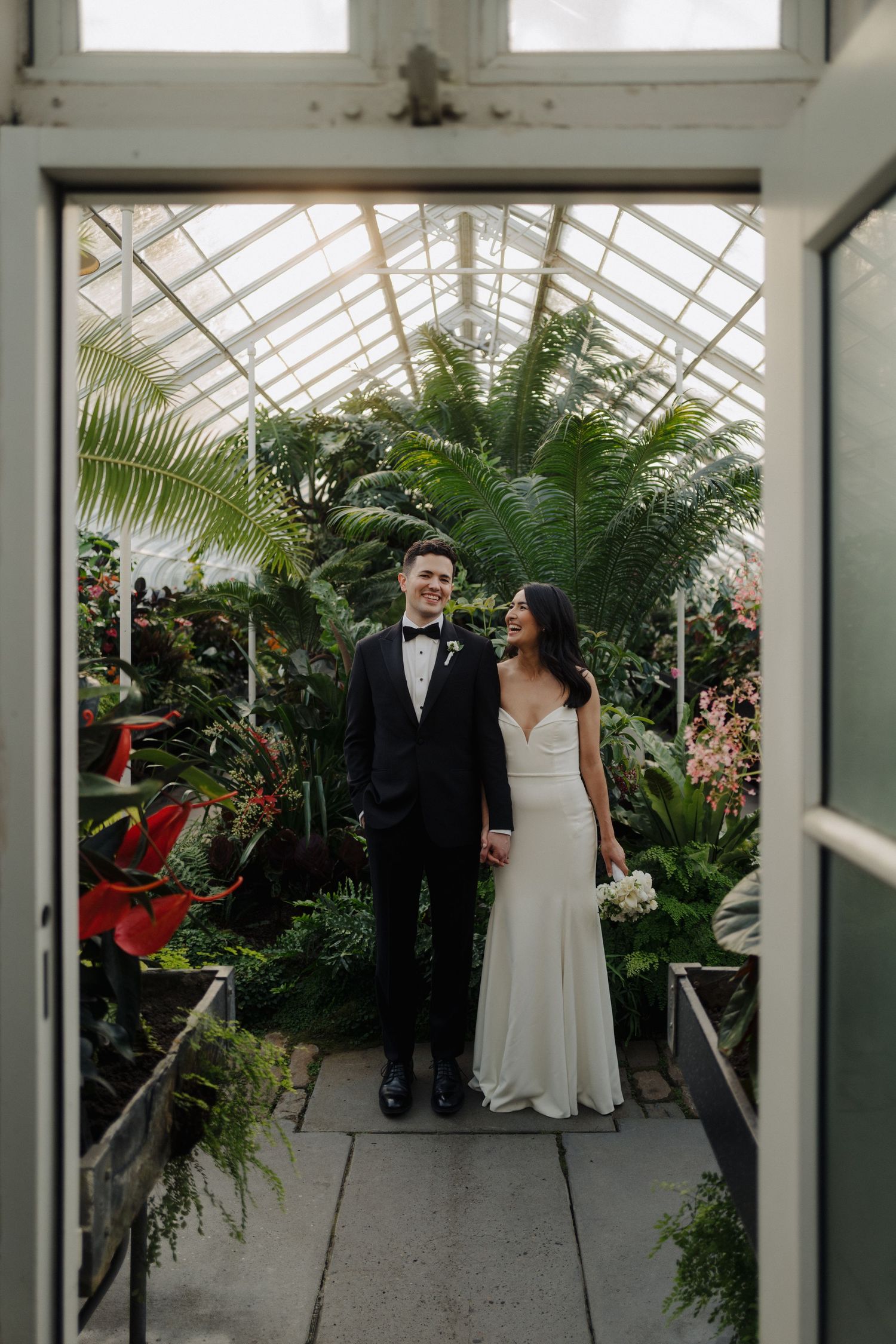 A wedding couple stands together in a greenhouse walkway surrounded by tropical greenery.