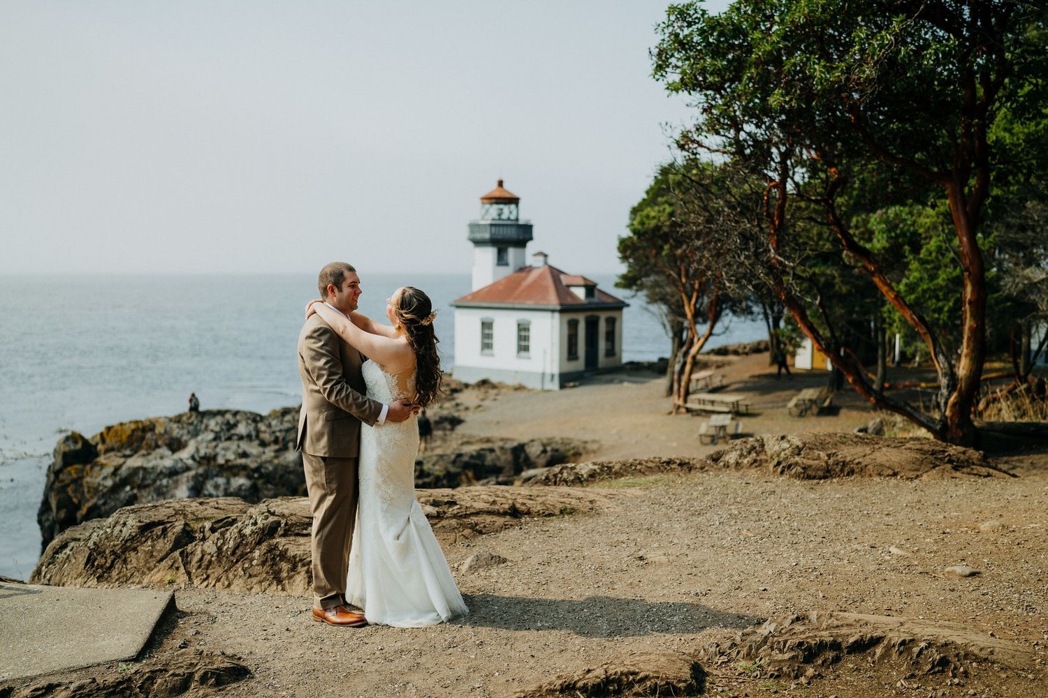 A couple stands together on rocky cliffs overlooking the ocean with a lighthouse in the background.