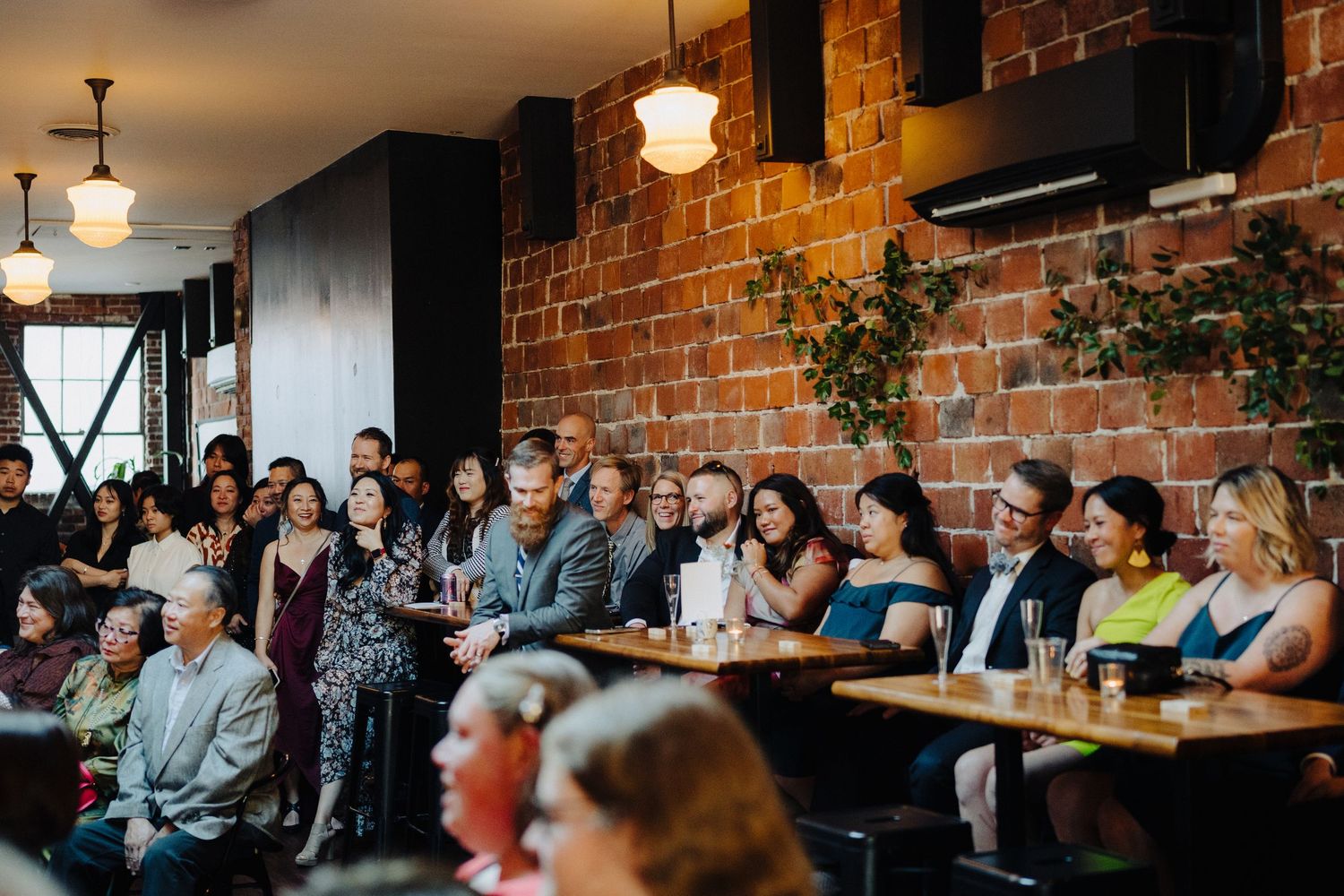 Wedding guests seated at long tables in a rustic brick-walled venue with pendant lighting.