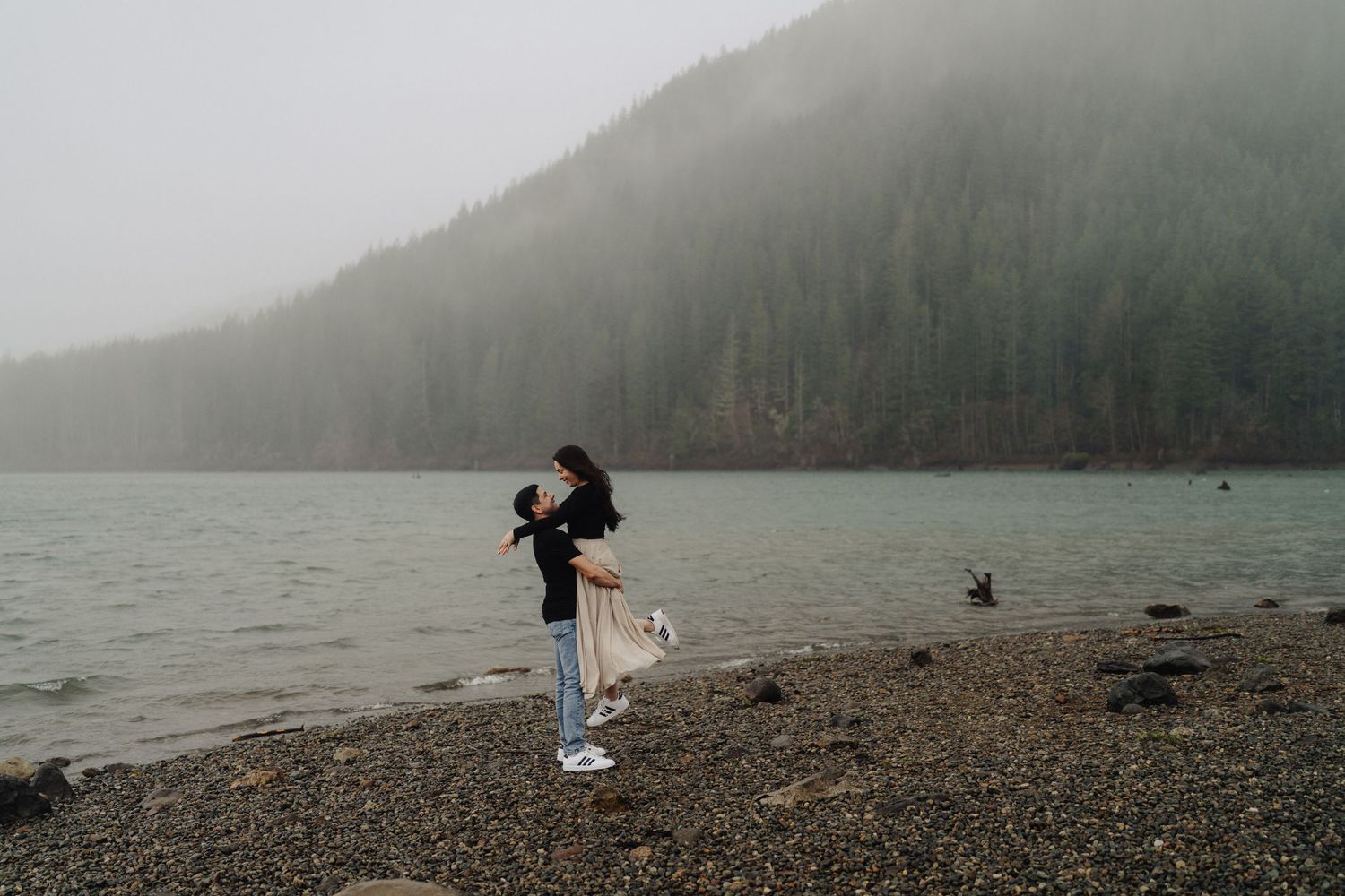 Playful couple spinning together on rocky lakeside with misty mountains in background.