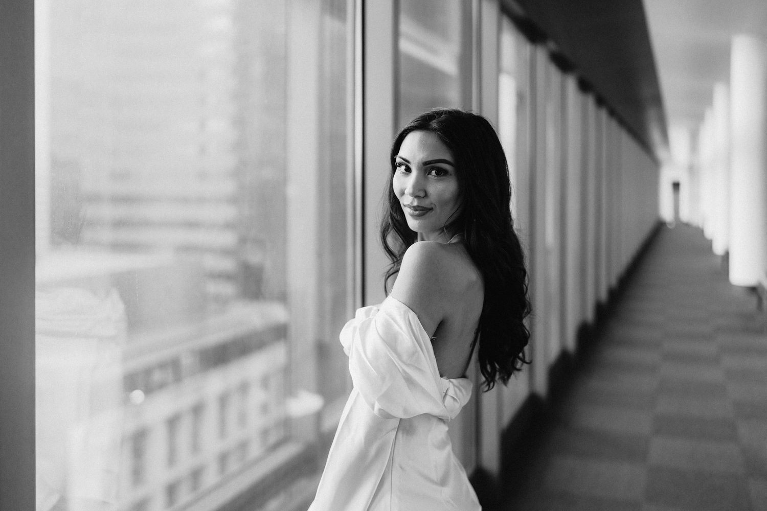A black and white portrait of a bride in an elegant off-shoulder dress posing by a window.
