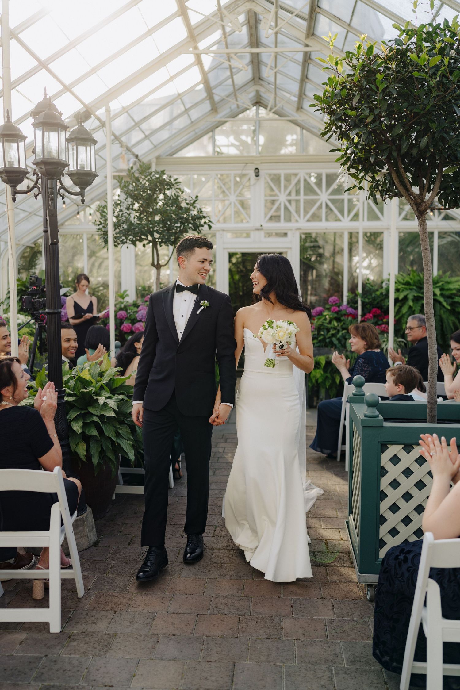 Newlyweds walking down the aisle in a greenhouse venue as guests applaud.