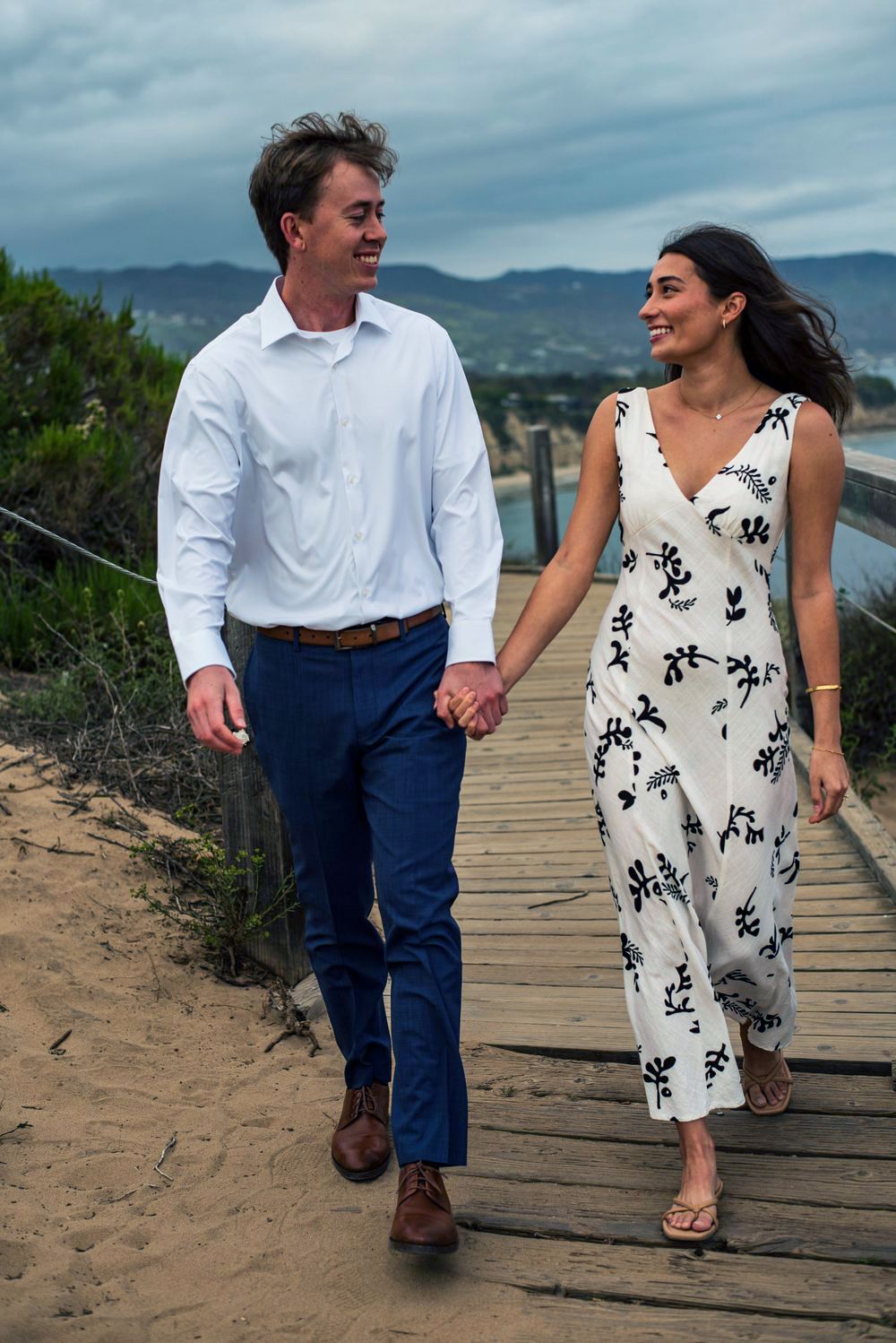 Couple walking hand in hand along a beachside boardwalk at sunset, smiling at each other during their engagement session in Malibu, CA