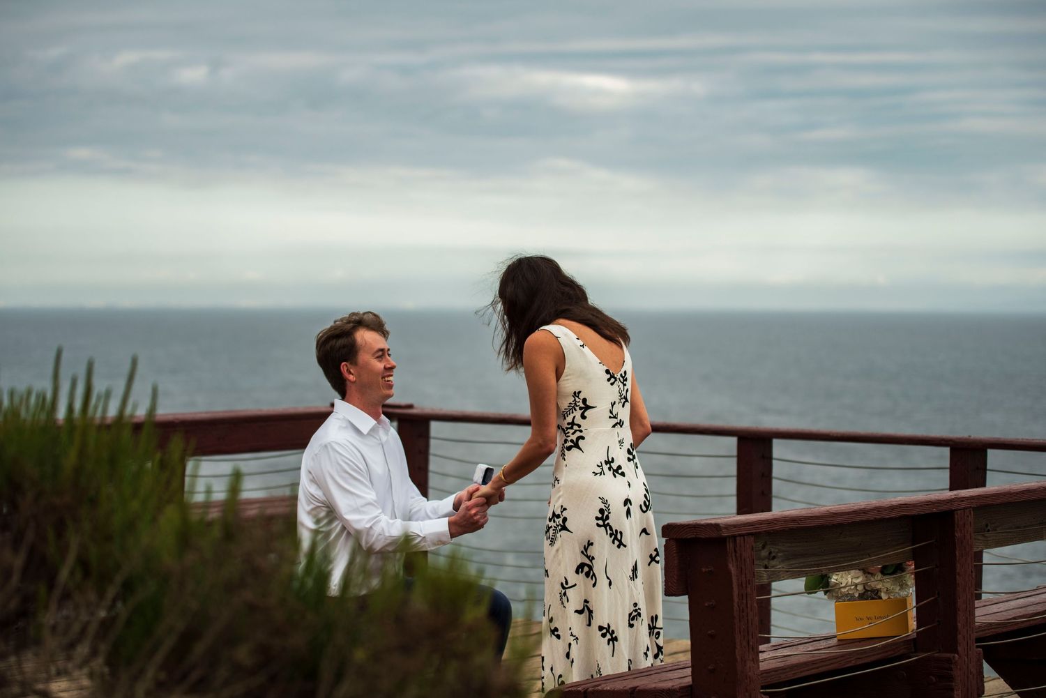 Man proposes on one knee to his partner on a scenic oceanfront deck in Malibu, California, during a heartfelt coastal engagement moment.