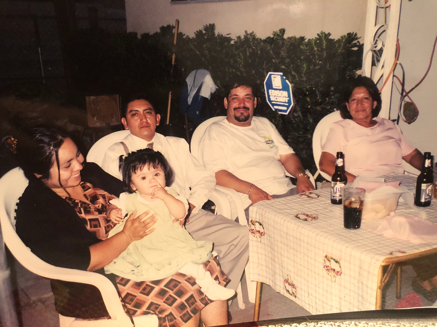 A family gathers around a white tablecloth-covered table outdoors with drinks and food during evening hours.