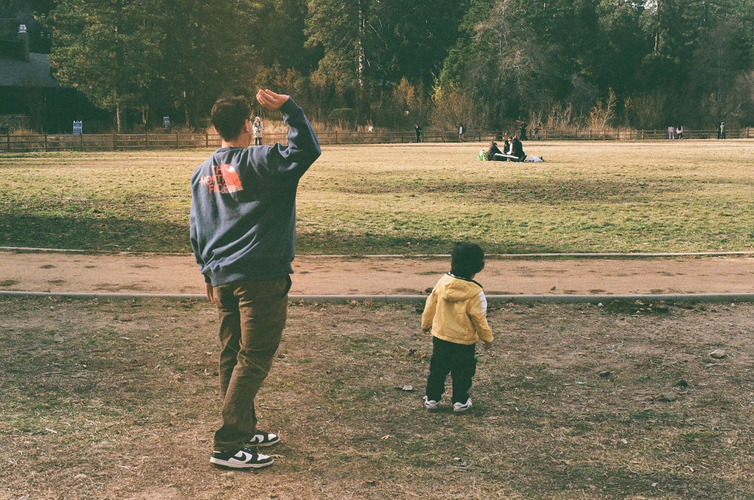 A figure in a hoodie and a small child in yellow stand together on a dirt path in a park on an autumn day.