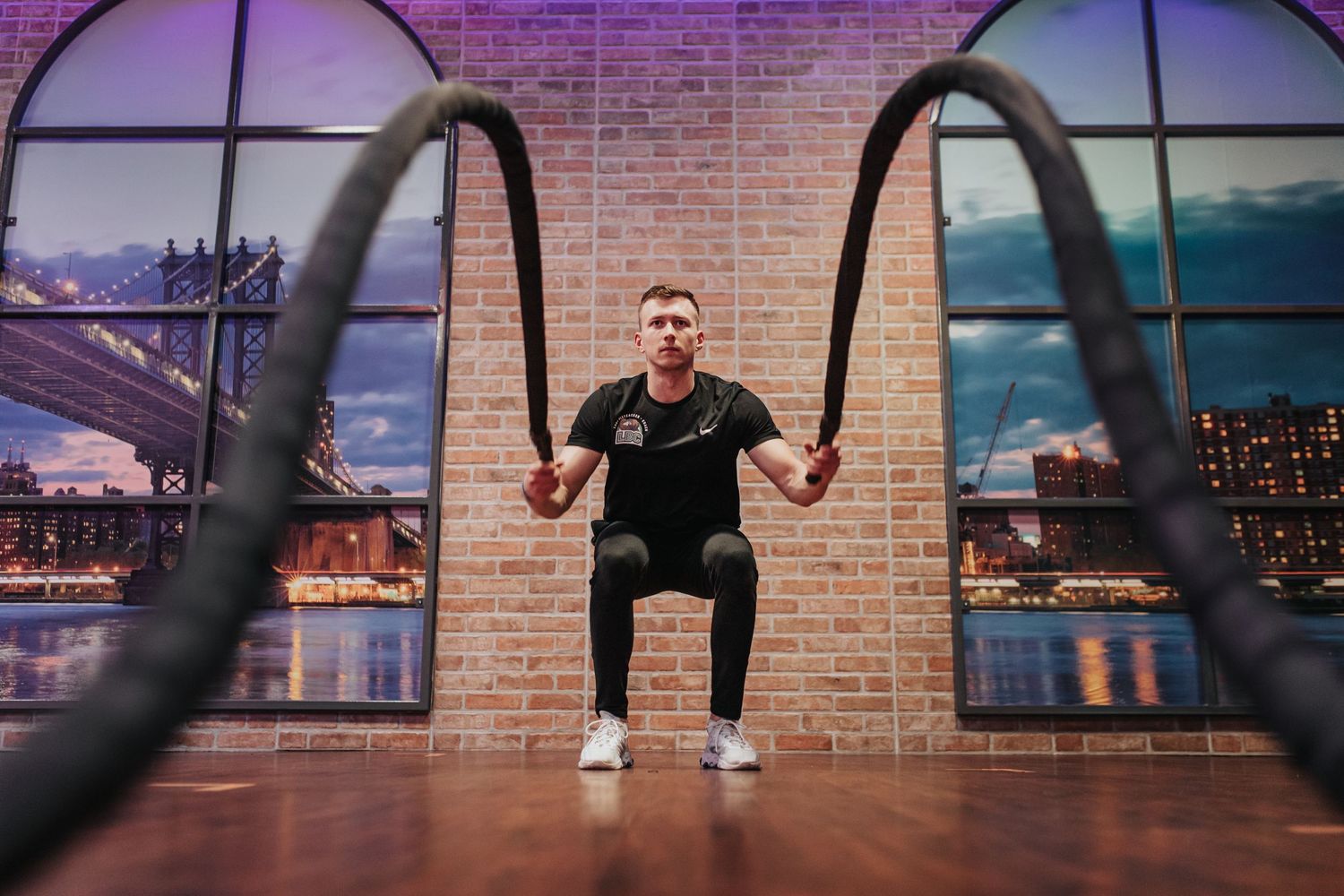Person working out with battle ropes in a modern gym with brick walls and floor-to-ceiling windows at sunset.