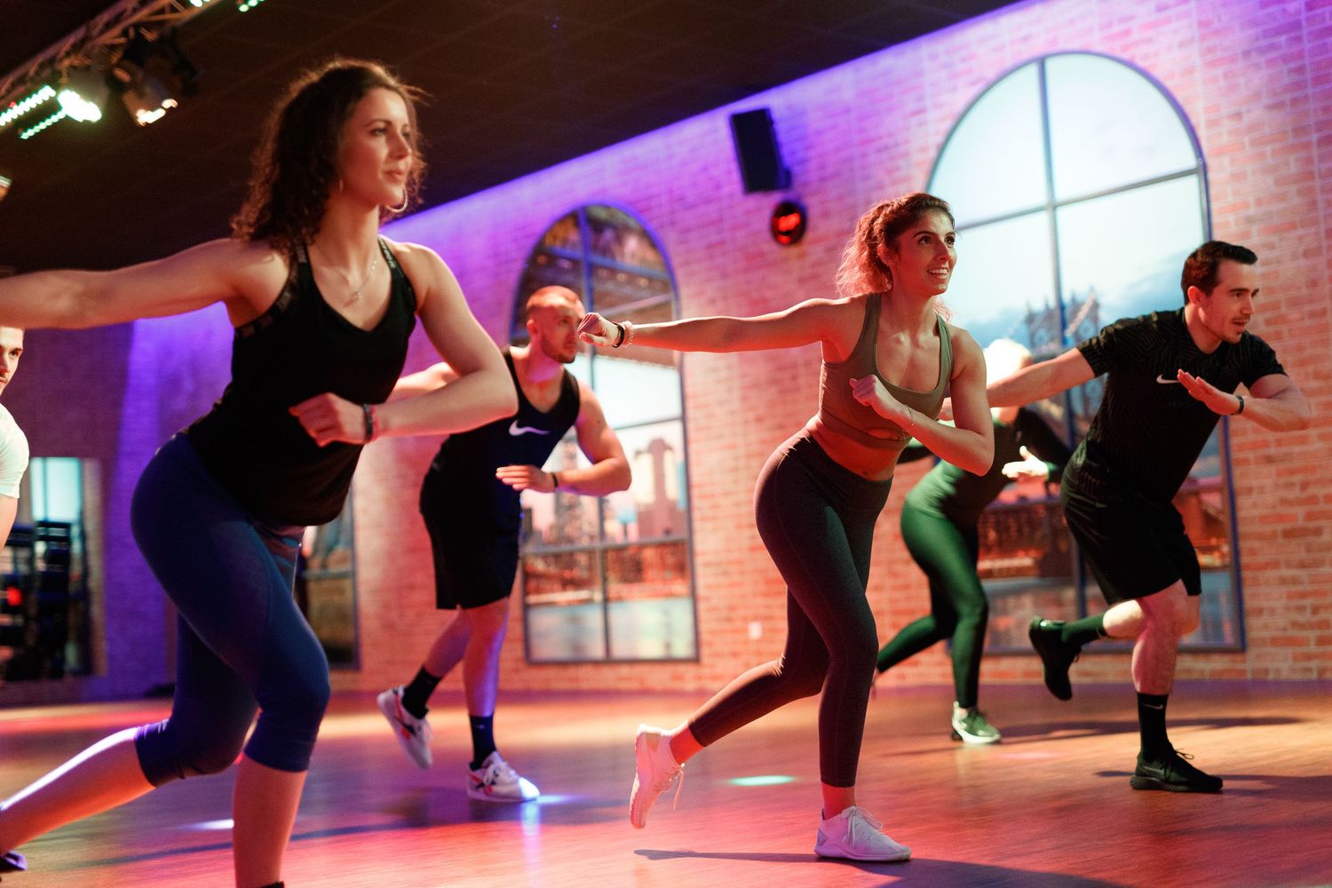 People participating in a high-energy group fitness class in a studio with purple lighting and arched windows.