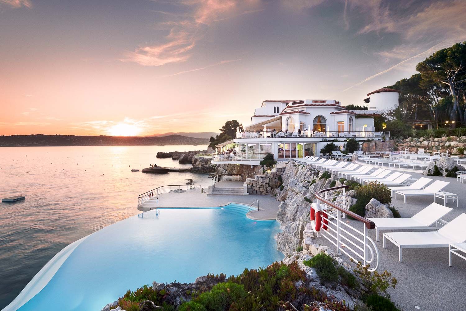 Luxury seaside resort with infinity pool overlooking Mediterranean waters during a pink-hued sunset.