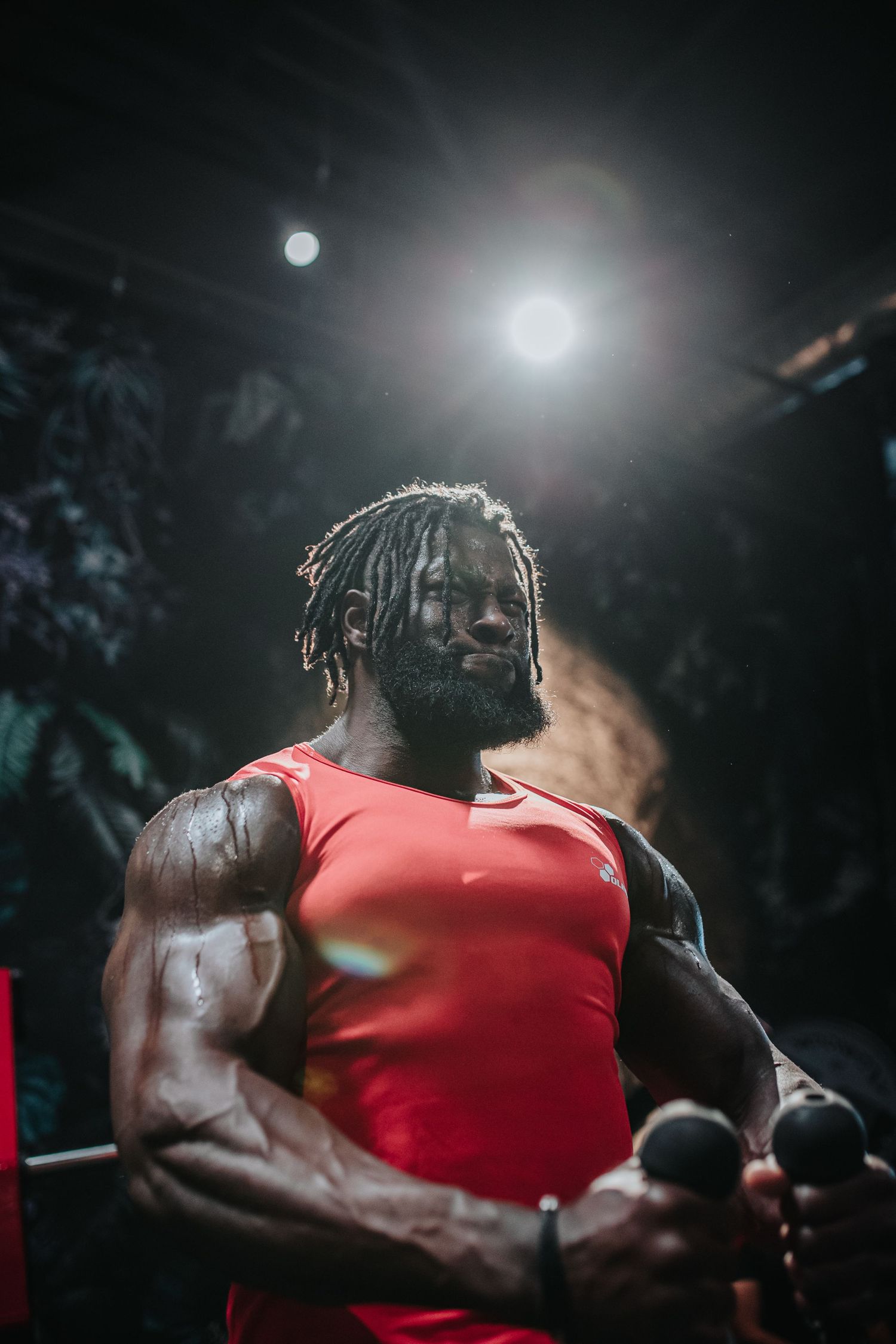 Athletic figure in red tank top performs intense back workout against dramatic dark background with spotlight effect.