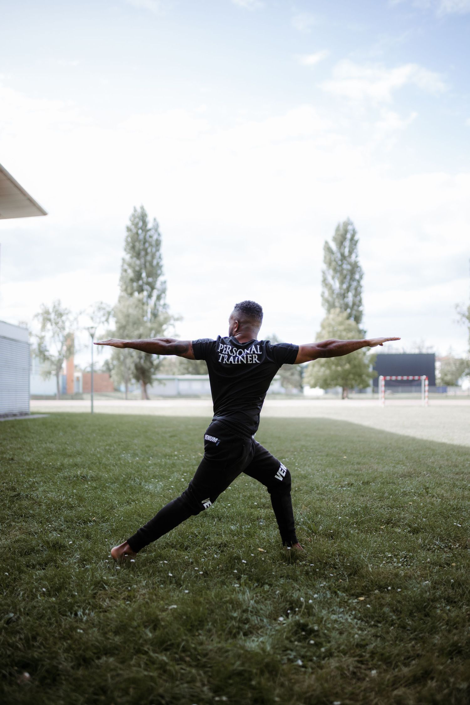 A person in black athletic wear performs a yoga warrior pose on a grassy field with trees in the background.