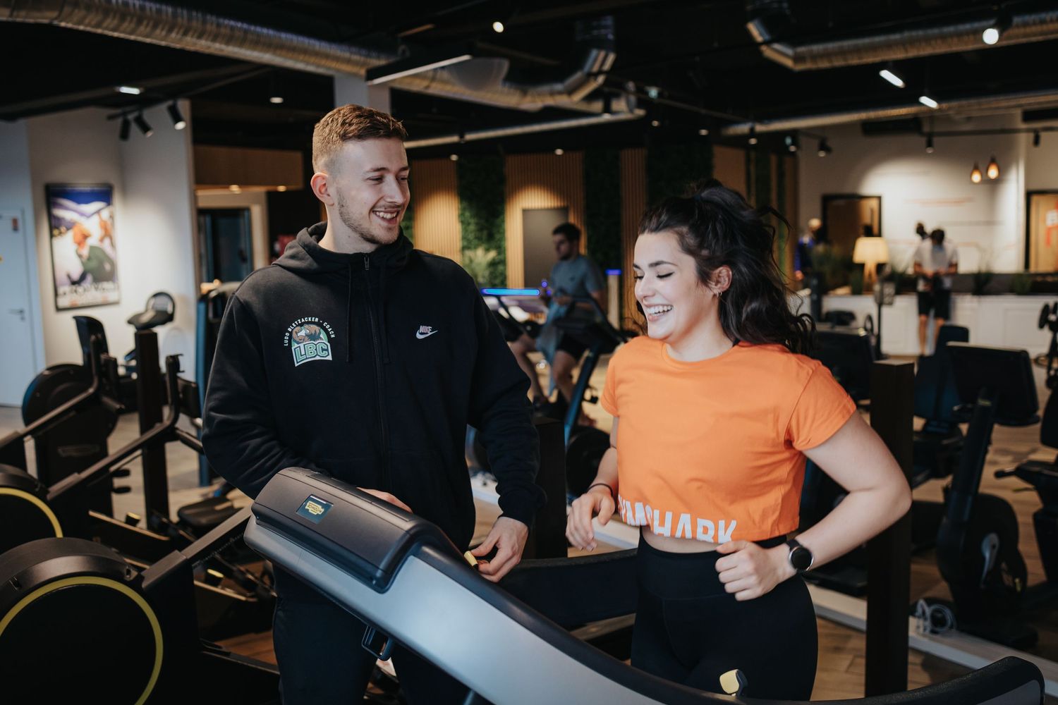 Two people work out in a fitness studio on weight machines with attentive staff guidance.