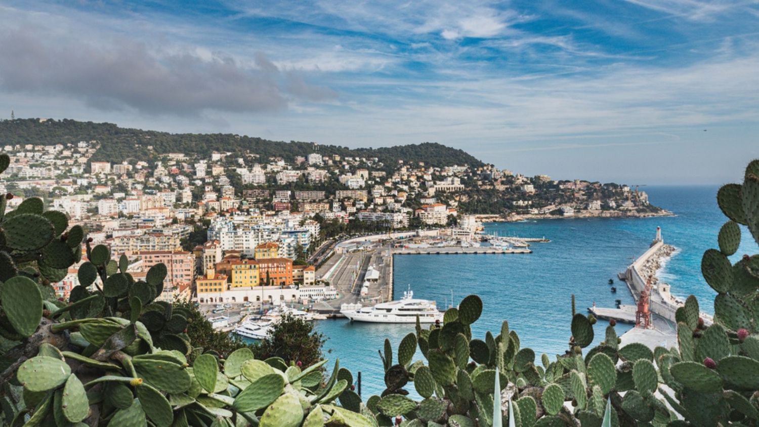 Aerial view of Nice's Mediterranean coastline with boats docked in harbor and hillside buildings under blue skies.