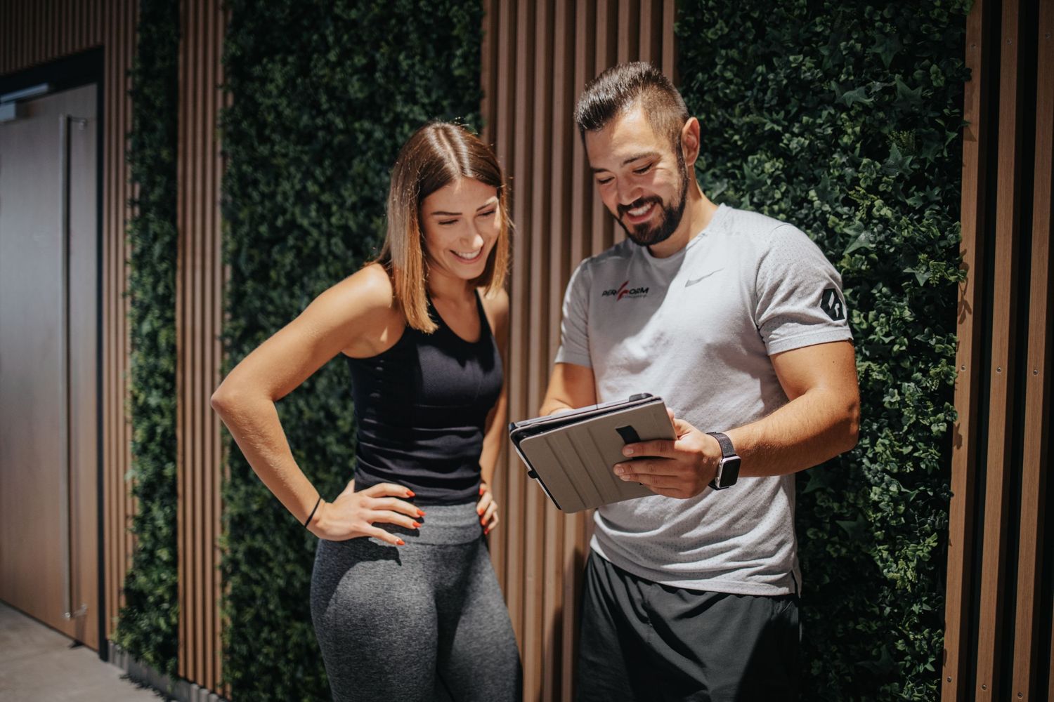 Two people in athletic wear reviewing fitness plans together on a tablet in front of a green plant wall.