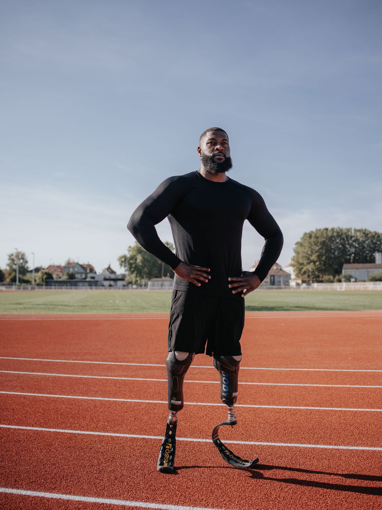 An athlete with prosthetic running blades stands confidently on a red running track on a sunny day.