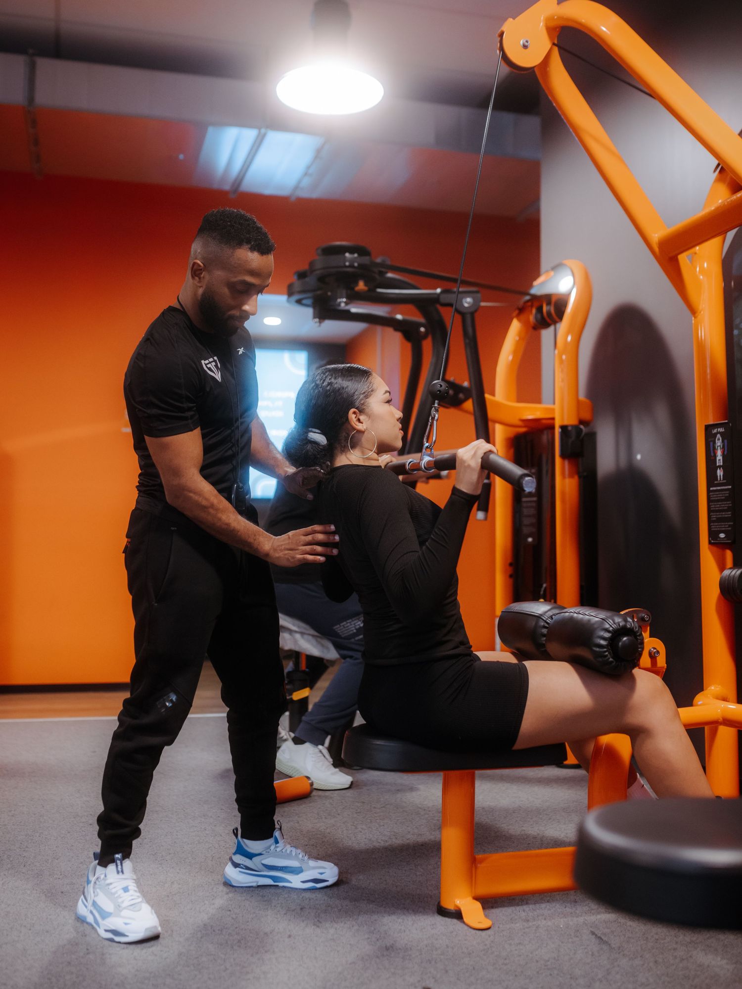 A trainer assists someone performing tricep exercises on an orange fitness machine in a modern gym setting.