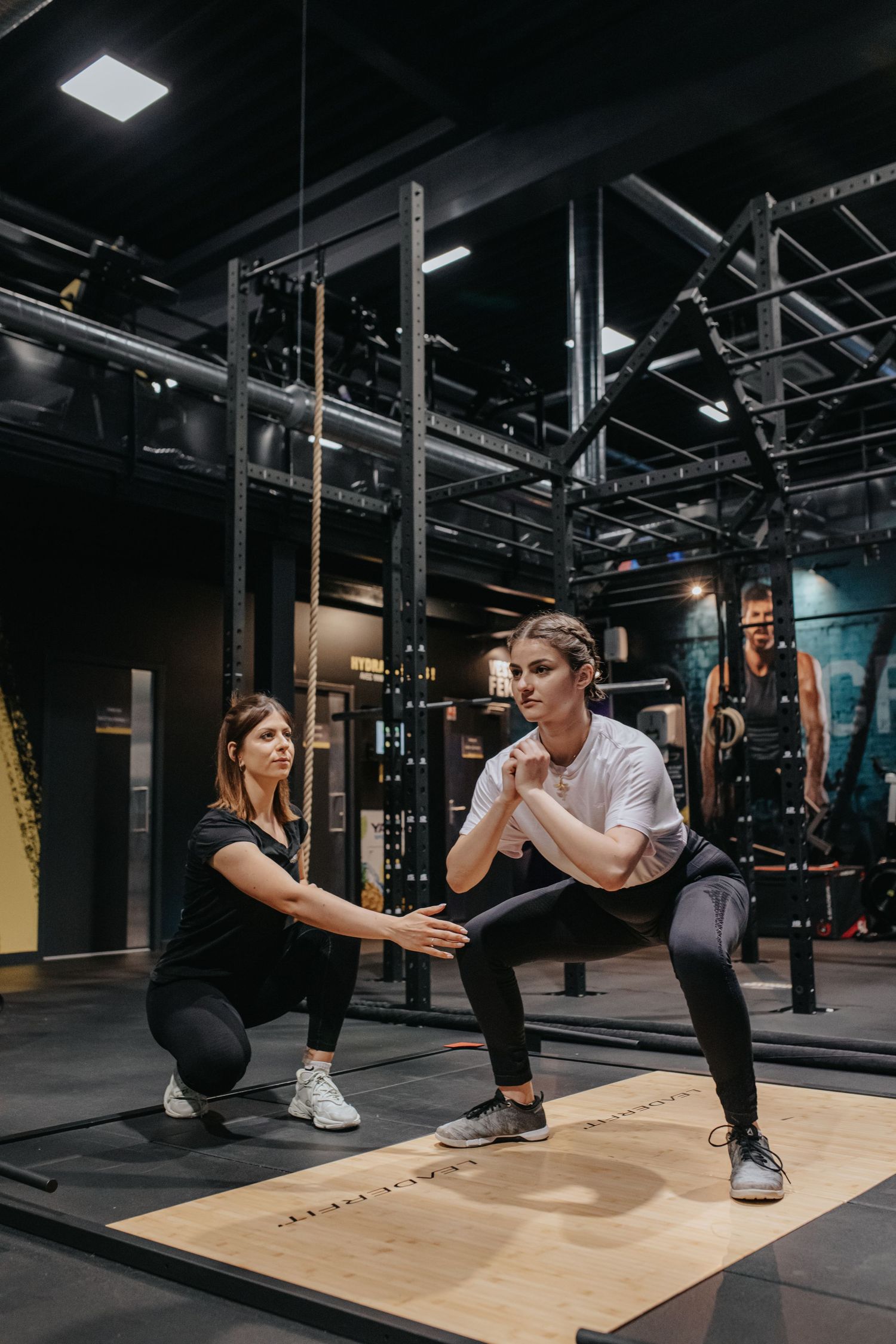 Personal trainer guiding client through proper squat form during strength training session in modern gym setting.