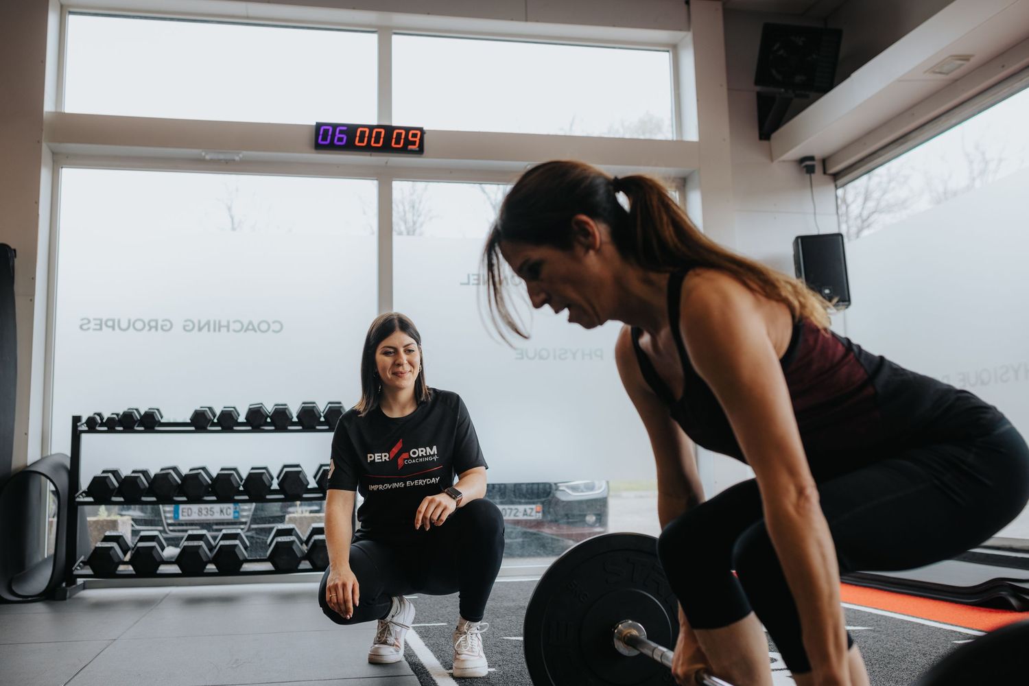 Two people work out in a gym with dumbbells and exercise equipment visible in the background.