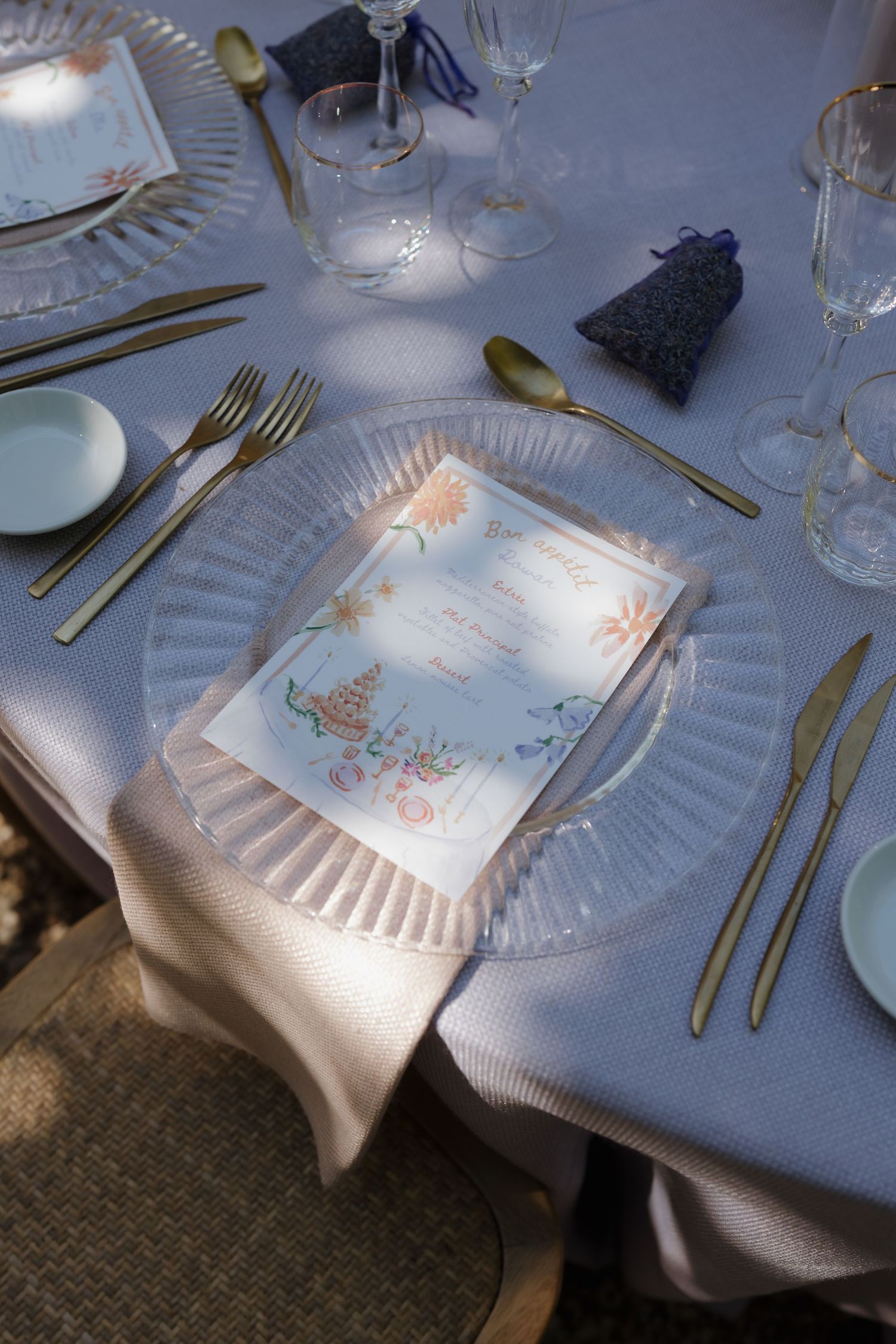 Elegant table setting with glass plates, golden cutlery, and delicate floral paper napkins on a light blue tablecloth.