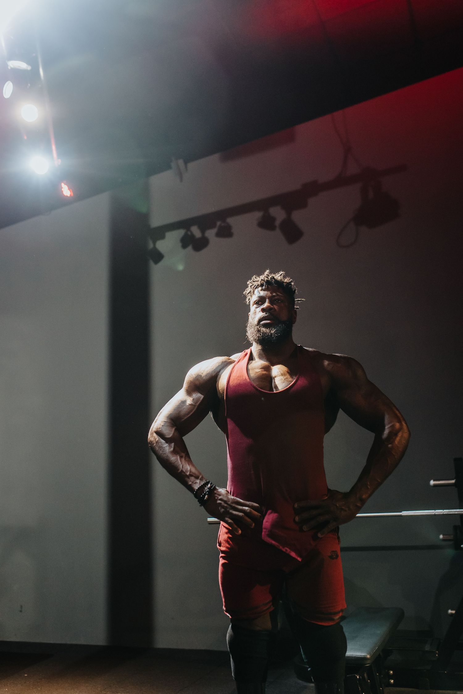 Muscular athlete in red activewear stands with hands on hips under dramatic stage lighting.