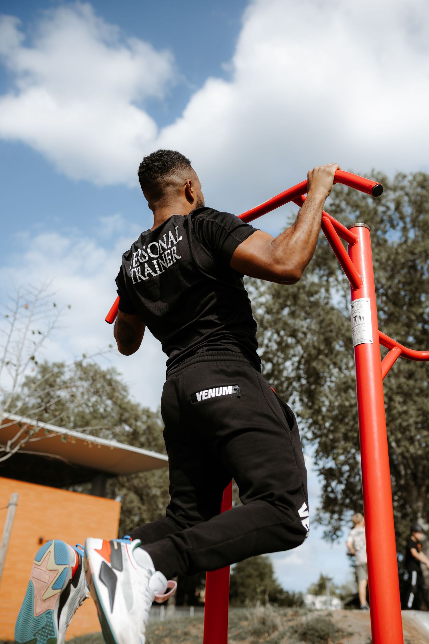 Person in black workout clothes performing pull-ups on a red outdoor exercise bar against a blue sky with clouds.