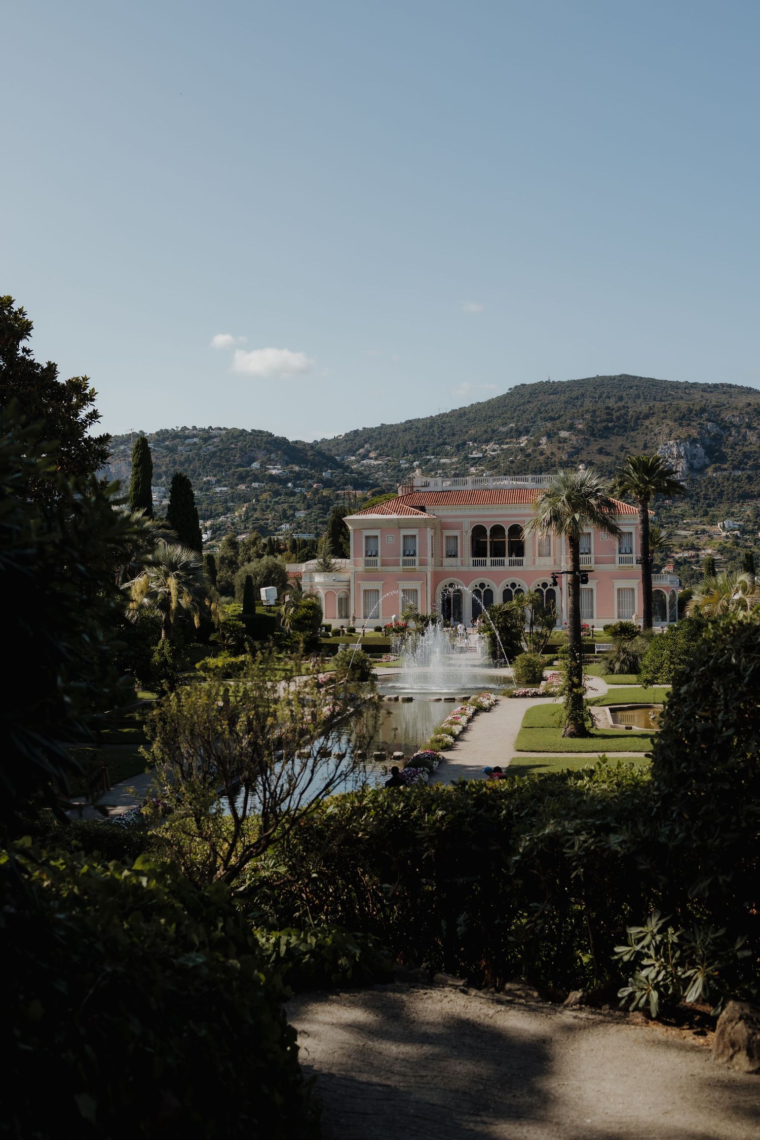 A pink Mediterranean villa with classical arches and formal gardens sits against a backdrop of rolling hills in the French Riviera.