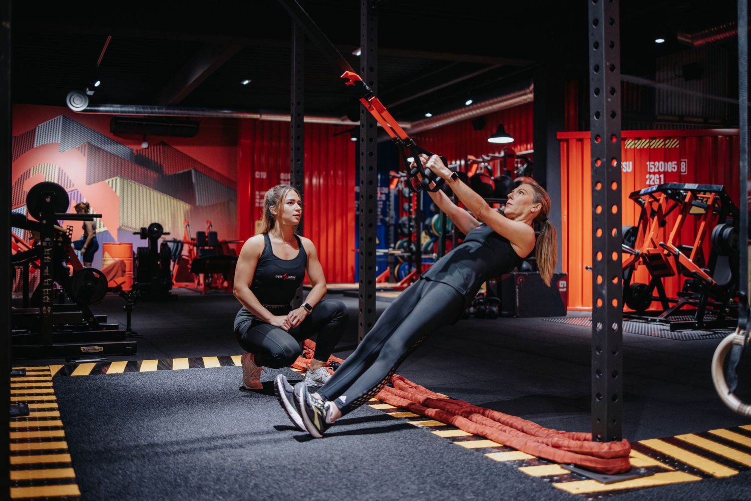 Six athletes perform dynamic full-body exercises across a red and black gym with modern fitness equipment.