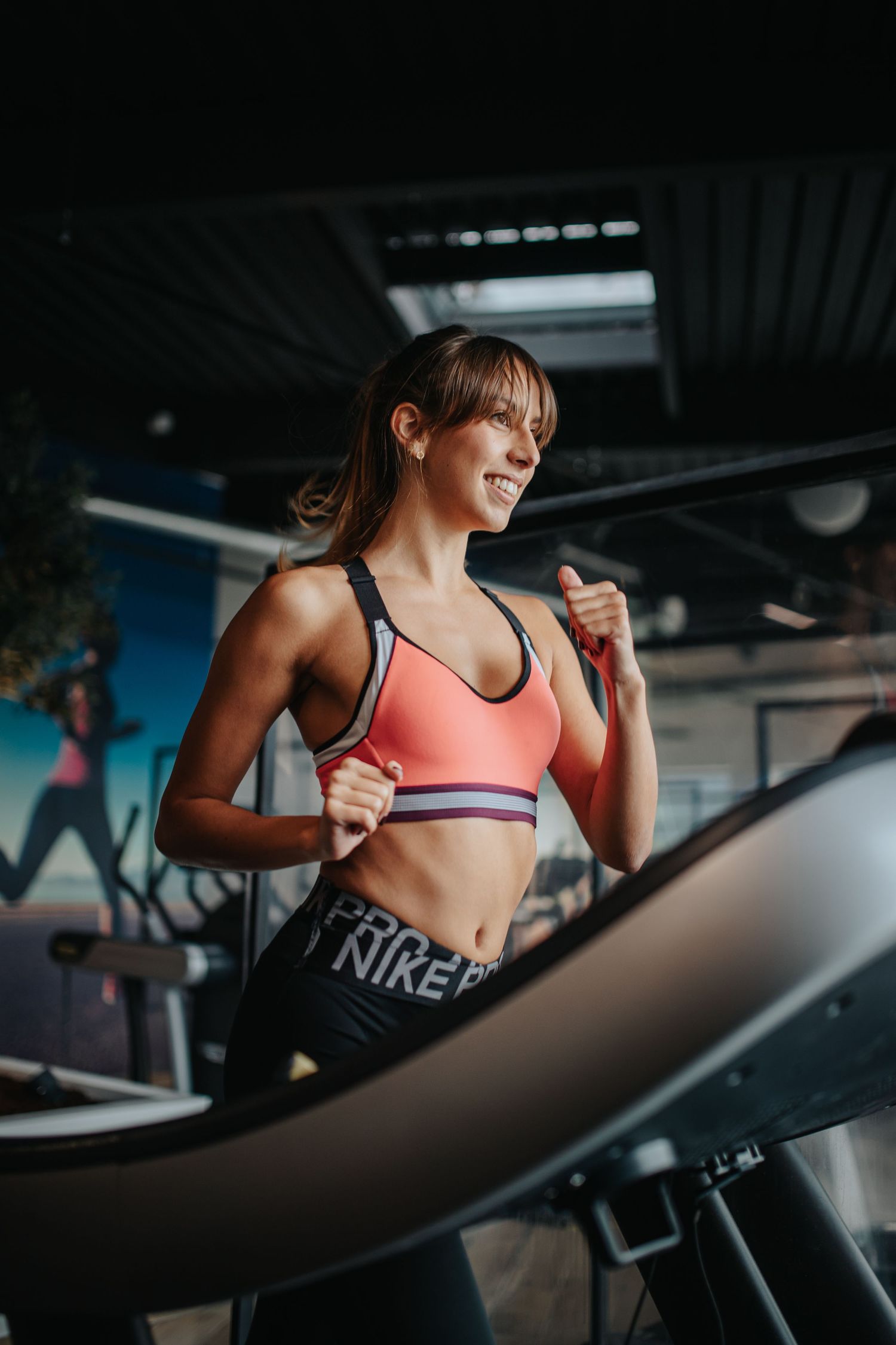 Person wearing a coral sports bra and Nike leggings works out on exercise equipment in a dimly lit gym.