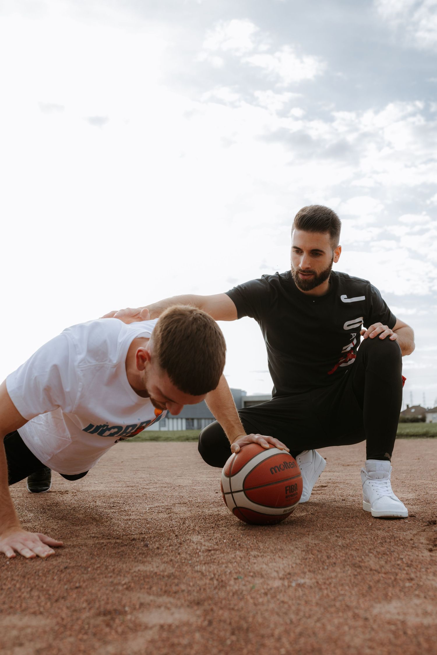 Two athletes perform one-arm pushups with a basketball on an outdoor basketball court at sunset.