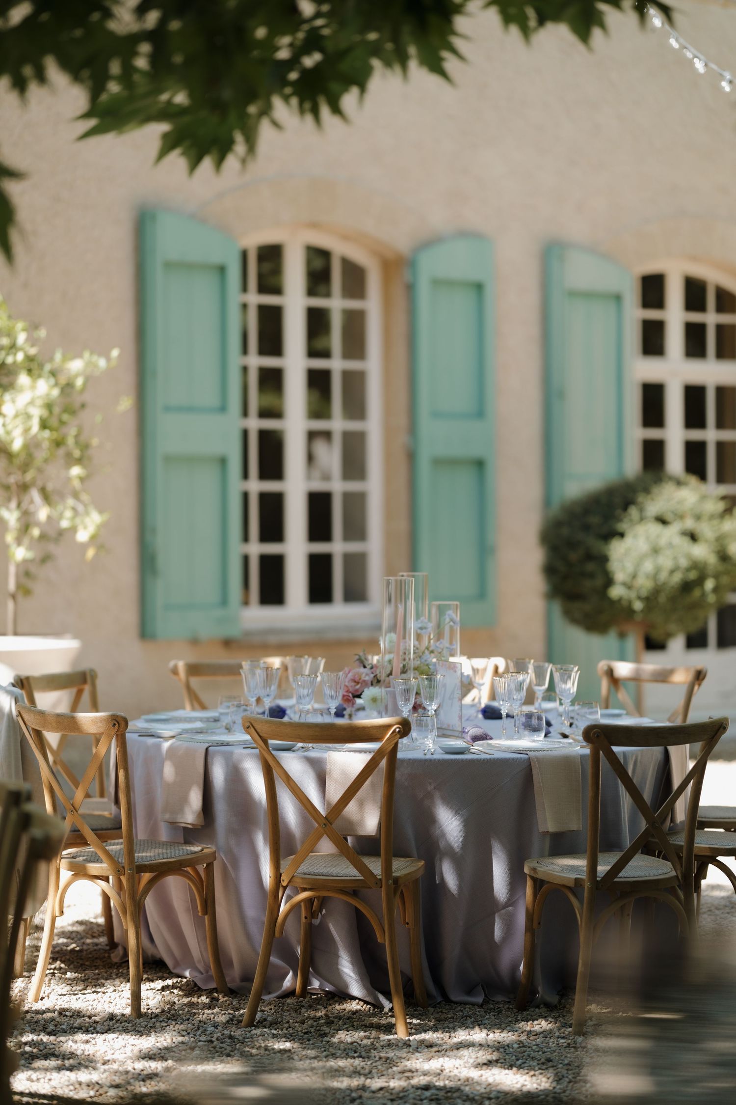 Elegant outdoor dining setup with wooden crossback chairs and gray tablecloths against a French cottage with mint shutters.
