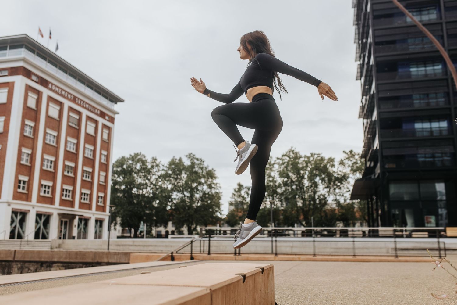A person performs a skateboarding trick in an urban plaza between brick and glass buildings at sunset.