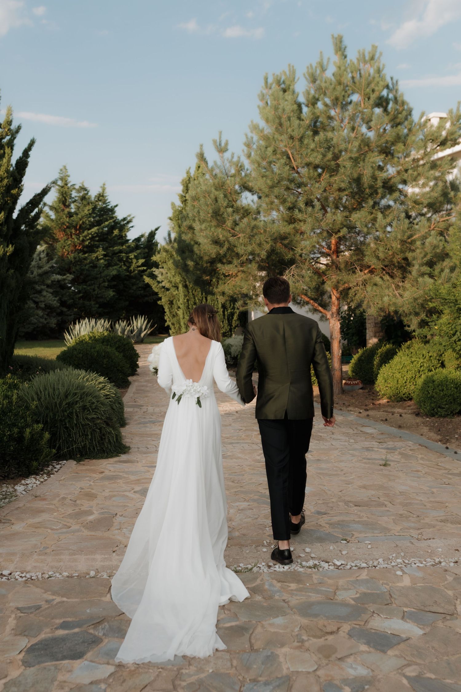 A couple walks along a stone path surrounded by pine trees and landscaping on their wedding day.